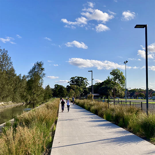 A path surrounded by bushland shrubs. A canal runs along the left and sporting courts along the right.